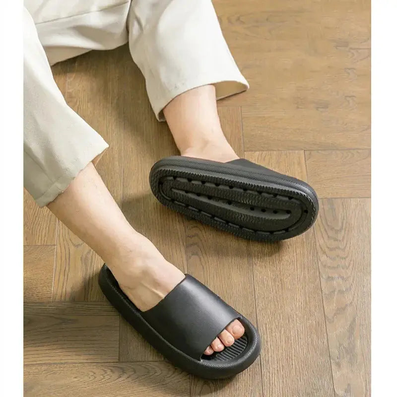 A person sits on a wooden floor in light pants, wearing CozySteps.uk’s Unisex EVA Beach & Bathroom Flip-Flops (US 6–12), with one sandal seen from above and the other showing its non-slip sole.