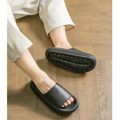 A person sits on a wooden floor in light pants, wearing CozySteps.uk’s Unisex EVA Beach & Bathroom Flip-Flops (US 6–12), with one sandal seen from above and the other showing its non-slip sole.